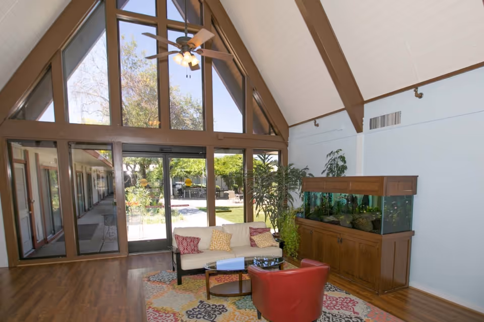 A bright living room area with a high vaulted ceiling featuring exposed wooden beams and a ceiling fan. The room has large floor-to-ceiling windows and glass doors that open to an outdoor garden area. Inside, there is a white sofa with colorful cushions, a red armchair, a glass coffee table on a patterned rug, and a large wooden fish tank with plants beside it.