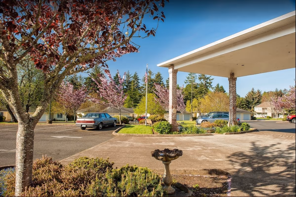 Covered porte-cochere entrance and driveway with parked cars, flowering trees, and landscaped grounds.