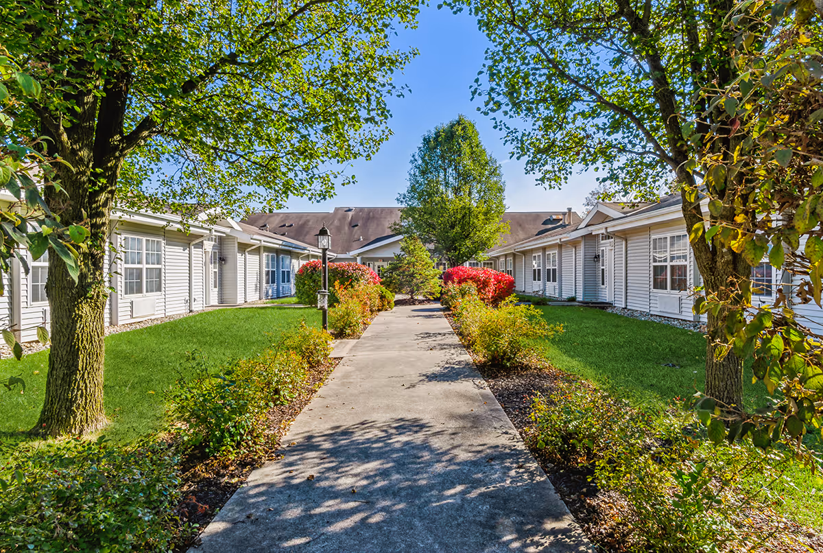 A sunny outdoor walkway lined with green grass, bushes, and trees leading to a single-story building with white siding and multiple windows under a clear blue sky.