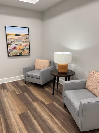A small seating area with two gray armchairs, each with a light pink cushion, positioned around a round dark wooden side table with a lamp on it. The floor has wood-patterned flooring, and a colorful landscape painting hangs on the light gray wall.