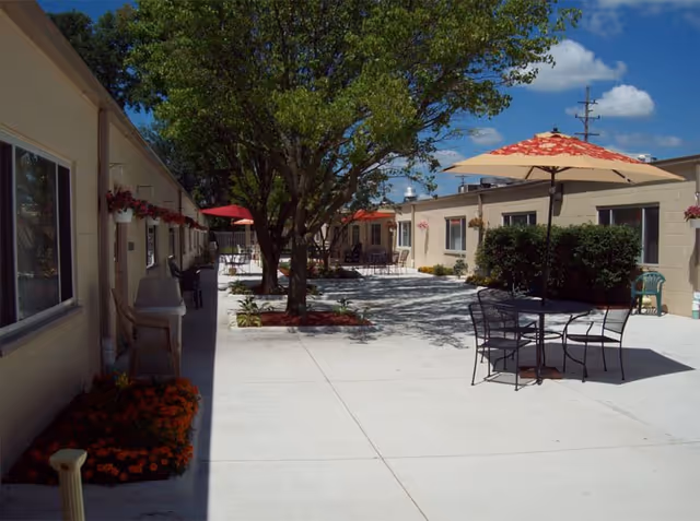 Outdoor courtyard area at CedarWoods Assisted Living & Memory Care with concrete walkways, trees, flower beds, patio tables with umbrellas, chairs, and beige single-story buildings under a blue sky with some clouds.