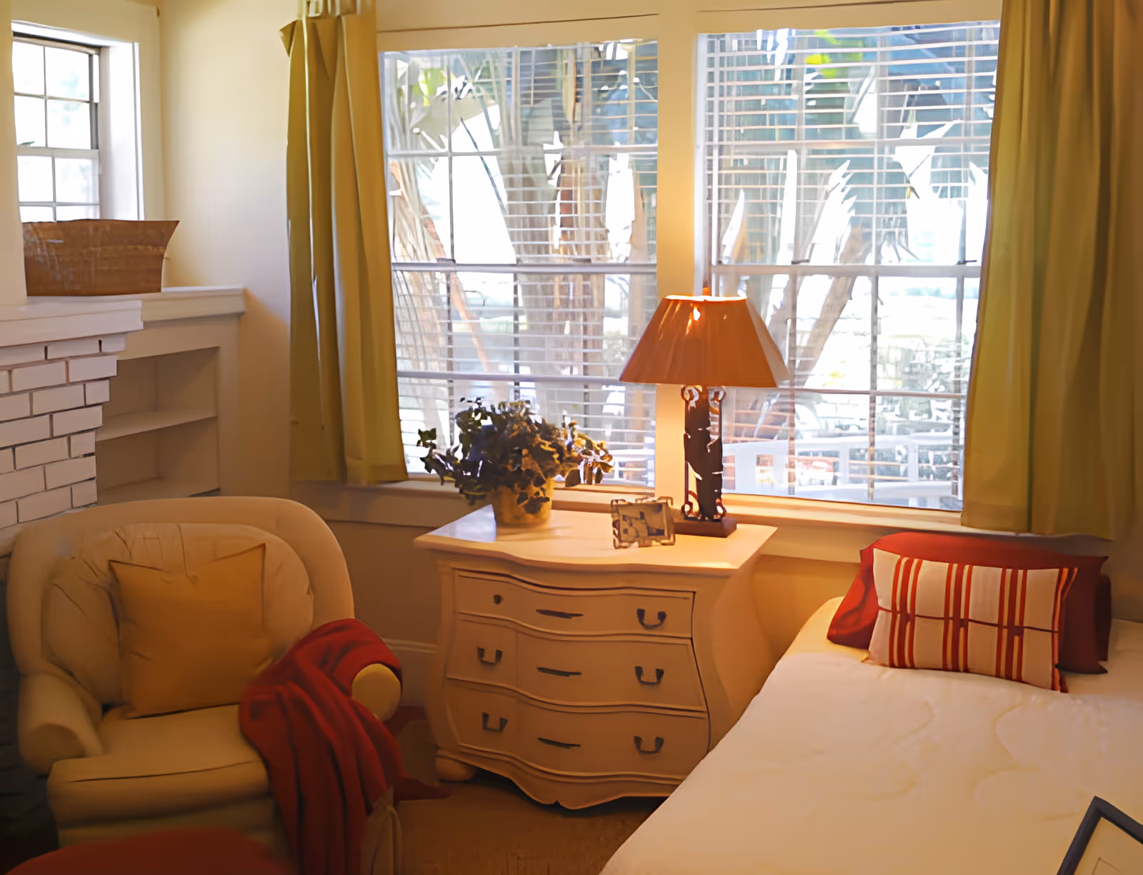 A cozy room with a cushioned armchair draped with a red blanket, a small white dresser with a table lamp and a potted plant on top, and a bed with decorative pillows. Large windows with yellow curtains let in natural light and show greenery outside.