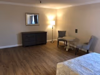 A simple bedroom with wooden flooring, a bed partially visible in the foreground, a dark dresser against the wall with a mirror above it, and a small round table with two upholstered chairs next to a floor lamp in the corner.