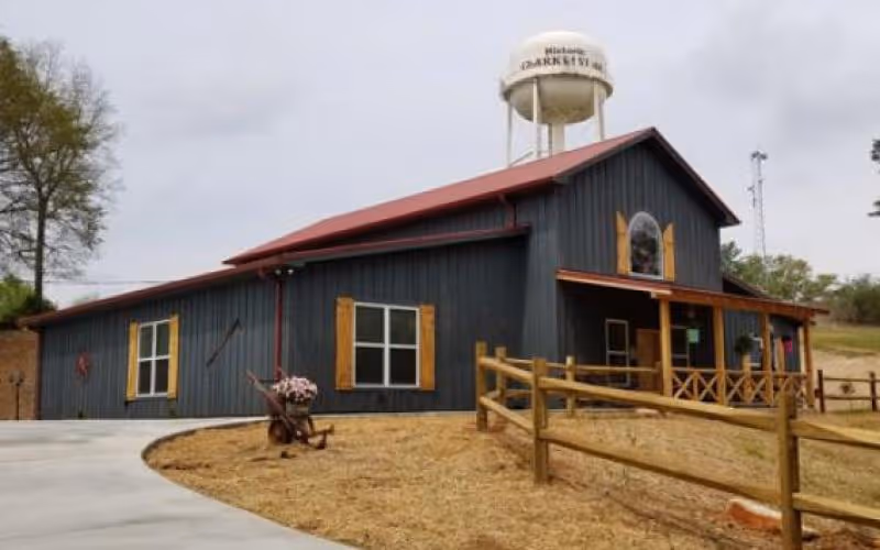 A dark-gray barn-style building with wooden shutters and a covered porch beside a water tower in a grassy lot.