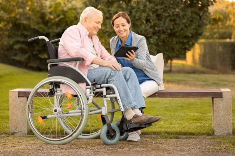 An elderly man in a wheelchair and a woman sitting on a bench outdoors, both looking at a tablet and smiling, with green grass and trees in the background.