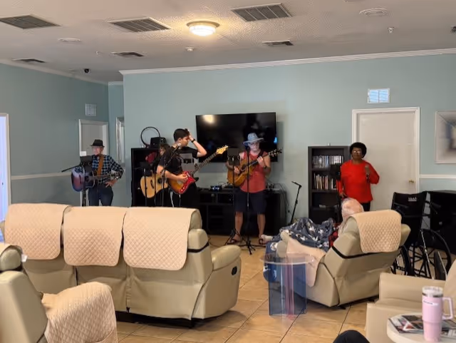 A group of four musicians performing in a living room area of a senior living facility. Three men are playing guitars and one woman is standing and singing. The room has beige recliners with quilted covers, a large flat-screen TV mounted on the wall, bookshelves, and a wheelchair visible on the right side.