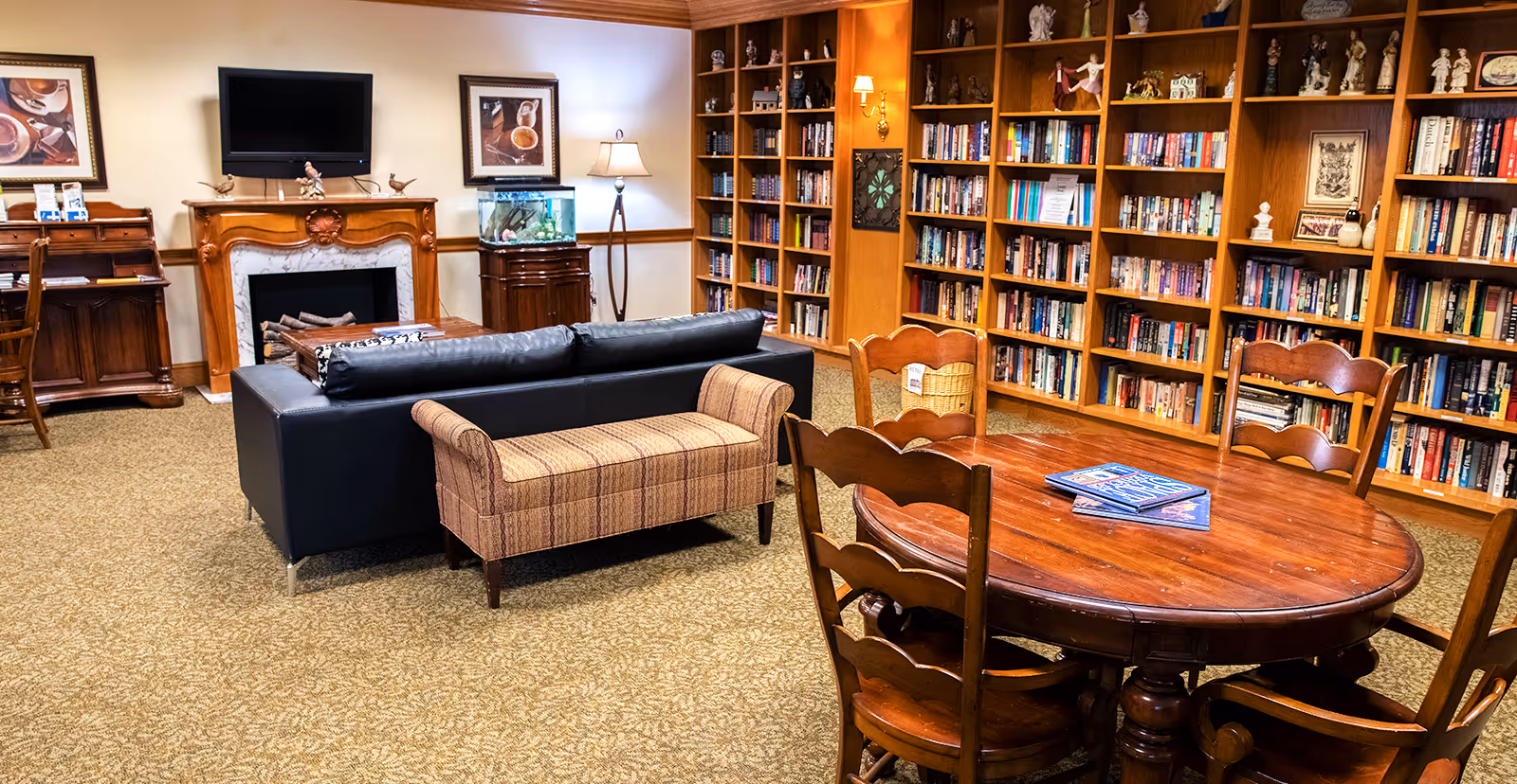 A cozy library and lounge area with wooden bookshelves filled with books and decorative items. There is a round wooden table with four chairs in the foreground, a black leather sofa with a patterned bench behind it, and a wooden fireplace with a TV mounted above it. The room has warm lighting and carpeted flooring.