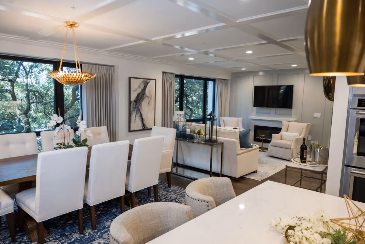 Open-plan dining and living area with a long dining table and upholstered chairs, a chandelier, sofas, and a wall-mounted TV.