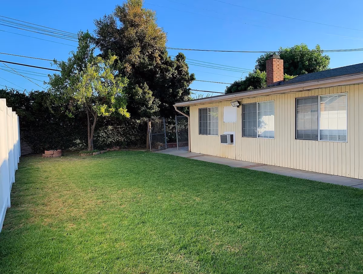 Grassy backyard with a single-story beige building showing windows and an air conditioner, a white fence, and trees under a clear blue sky.