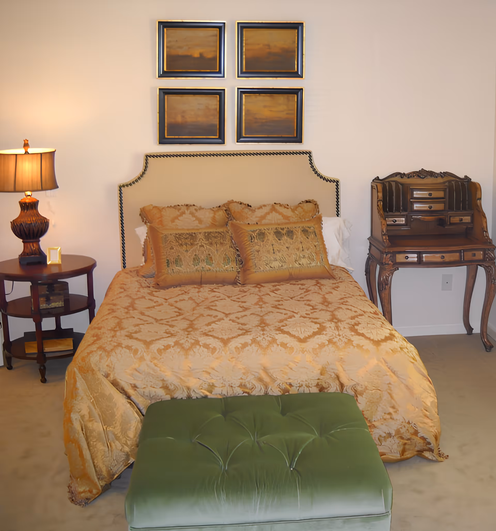 A neatly made bedroom with a gold patterned bed, green tufted ottoman at the foot, a lamp on a round nightstand to the left and an ornate wooden desk to the right under four framed pictures.