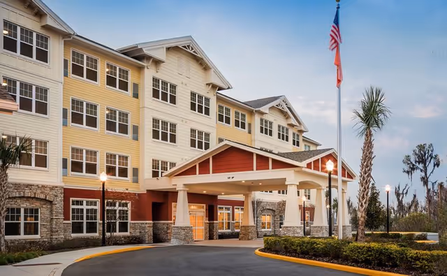 Exterior view of a multi-story senior living facility building with a covered entrance, stone and siding facade, multiple windows, palm trees, and two flagpoles displaying the American flag and another flag.