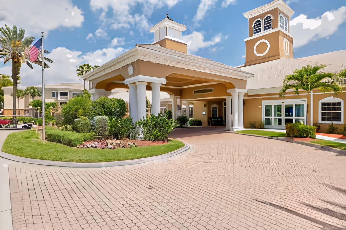 Entrance of a senior living facility with a covered driveway, beige building with white columns, manicured bushes and trees, and a clear blue sky with some clouds.