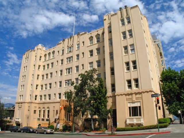 Exterior view of a large beige multi-story building under a partly cloudy sky with trees and parked cars in front.