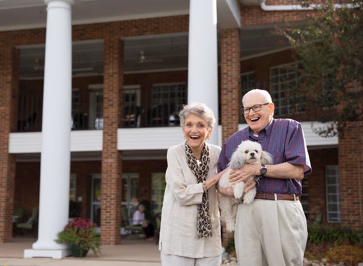 An elderly couple standing outside a brick building with white columns. The man is holding a small white dog, and both are smiling and appear happy. There are plants and outdoor seating visible in the background.