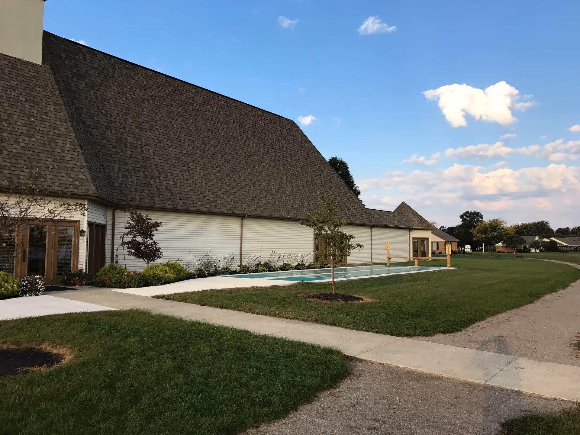 Exterior view of a single-story building with a steep shingled roof, lawn, sidewalk, and a small reflecting pool under a blue sky.