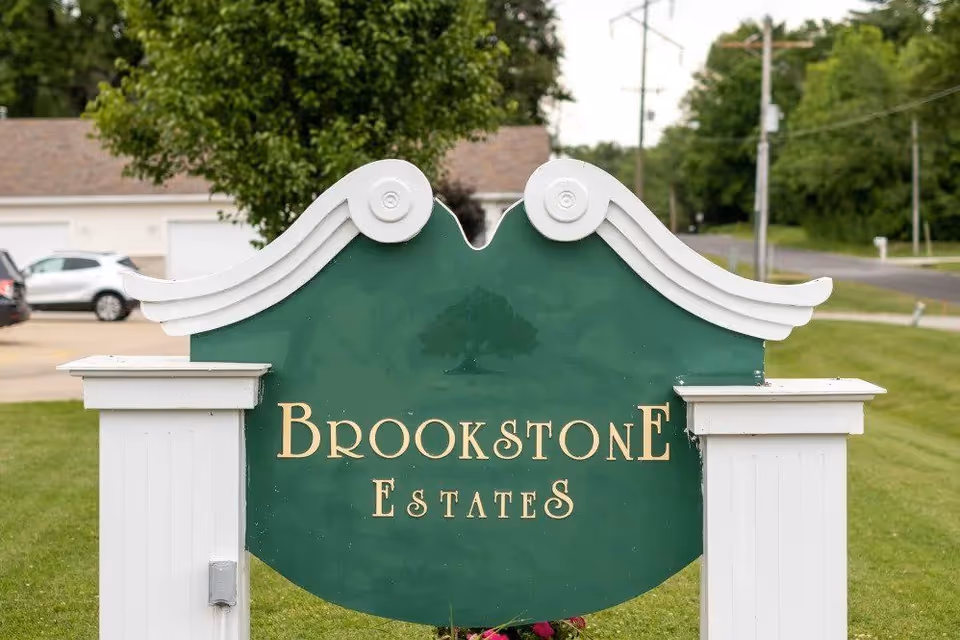 A green and white decorative sign with the text 'Brookstone Estates' displayed outdoors on a grassy area near a road, with trees and parked cars in the background.