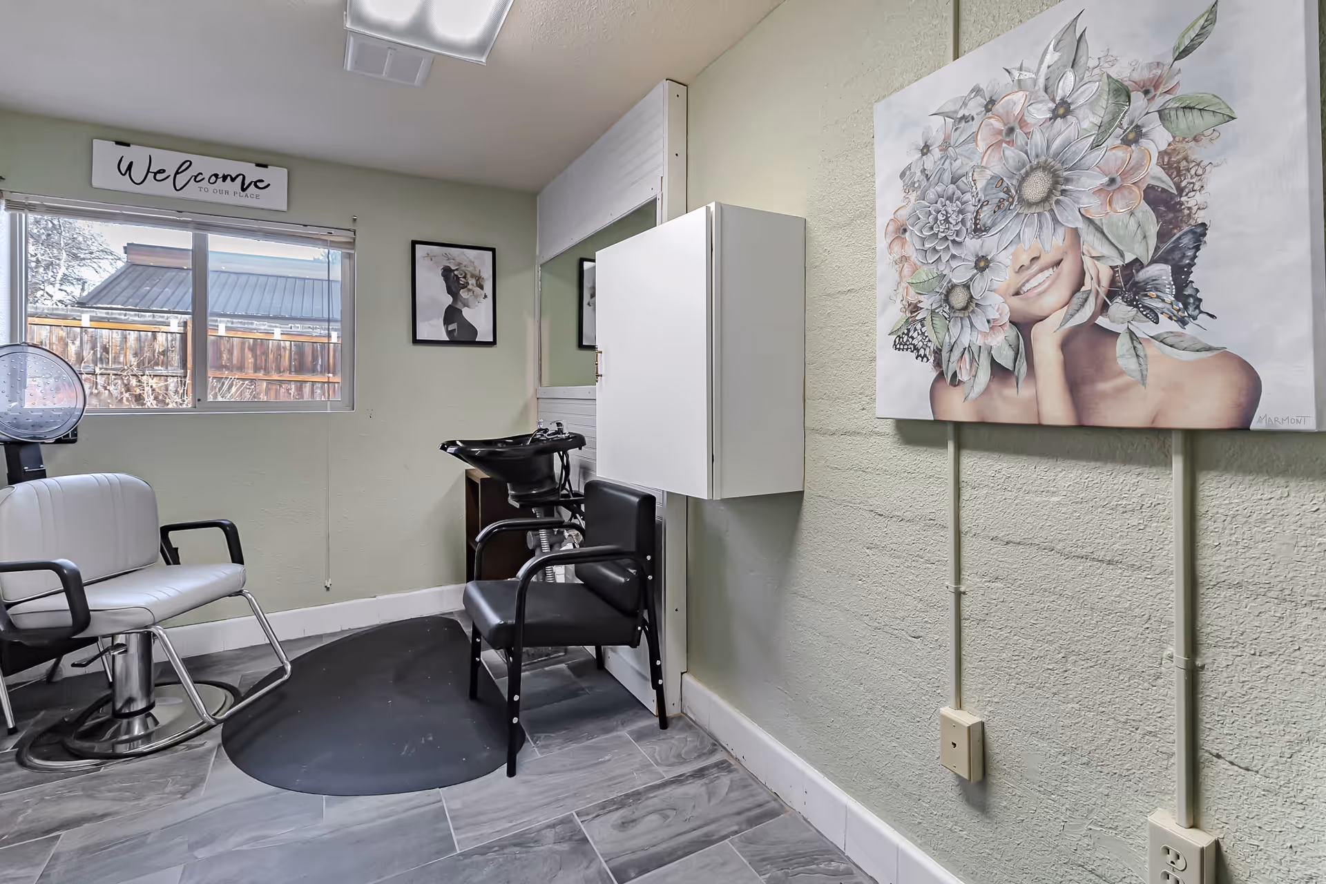 A small salon area with a white styling chair, a black shampoo chair with a wash basin, a large mirror, and a window with a 'Welcome to our place' sign above it. The walls are light green with a textured finish, and there is a colorful painting of a woman with flowers and butterflies on her head hanging on the right wall.
