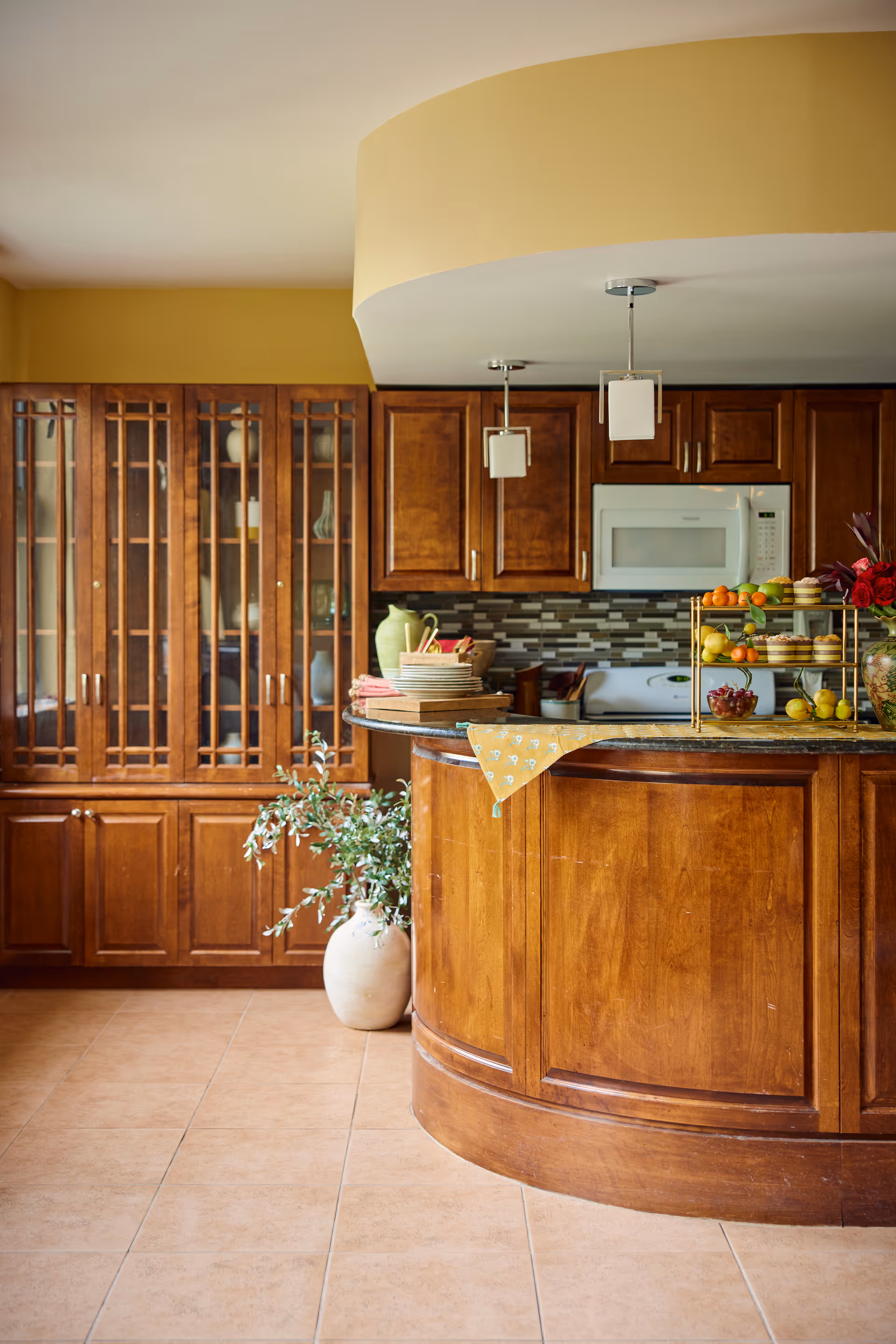 A warm kitchen interior featuring wooden cabinets and a curved wooden island with a granite countertop. The kitchen has a tiled backsplash, a white microwave, and a stove. On the island, there are plates, a tiered fruit stand with various fruits, and a vase with flowers. A large ceramic pot with a green plant is placed on the floor next to the island. The walls are painted yellow, and the floor is tiled.