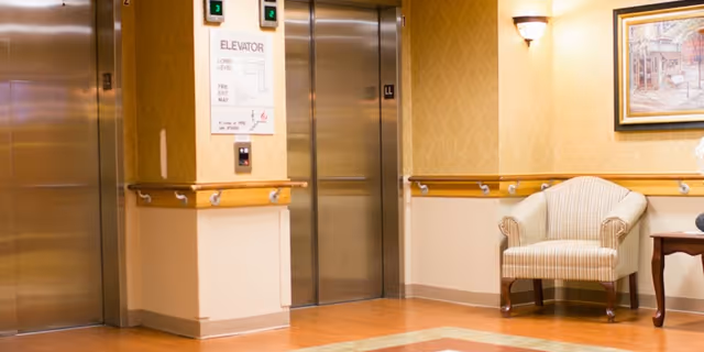 Interior view of a hallway in a senior living facility featuring two closed stainless steel elevator doors. The walls are decorated with a patterned wallpaper and wooden chair rails. A striped upholstered armchair is placed next to a wooden side table with a floral arrangement. A framed painting and a wall sconce light fixture are mounted on the wall above the chair.