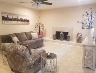 A cozy living room with a patterned armchair and a gray sofa adorned with pillows. There is a small round side table in front of the armchair. The room features a fireplace with decorative lanterns and a large vase filled with decorative balls. A ceiling fan with lights is mounted on the ceiling, and a large piece of artwork hangs on the wall above the sofa. The floor is tiled in a light color.