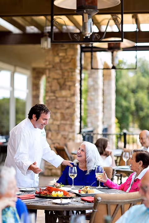 A chef in a white uniform is engaging warmly with elderly women seated at an outdoor dining table. The table is set with plates of food, including seafood and garnishes, and glasses of white wine. The setting is a covered patio with stone pillars and greenery in the background.