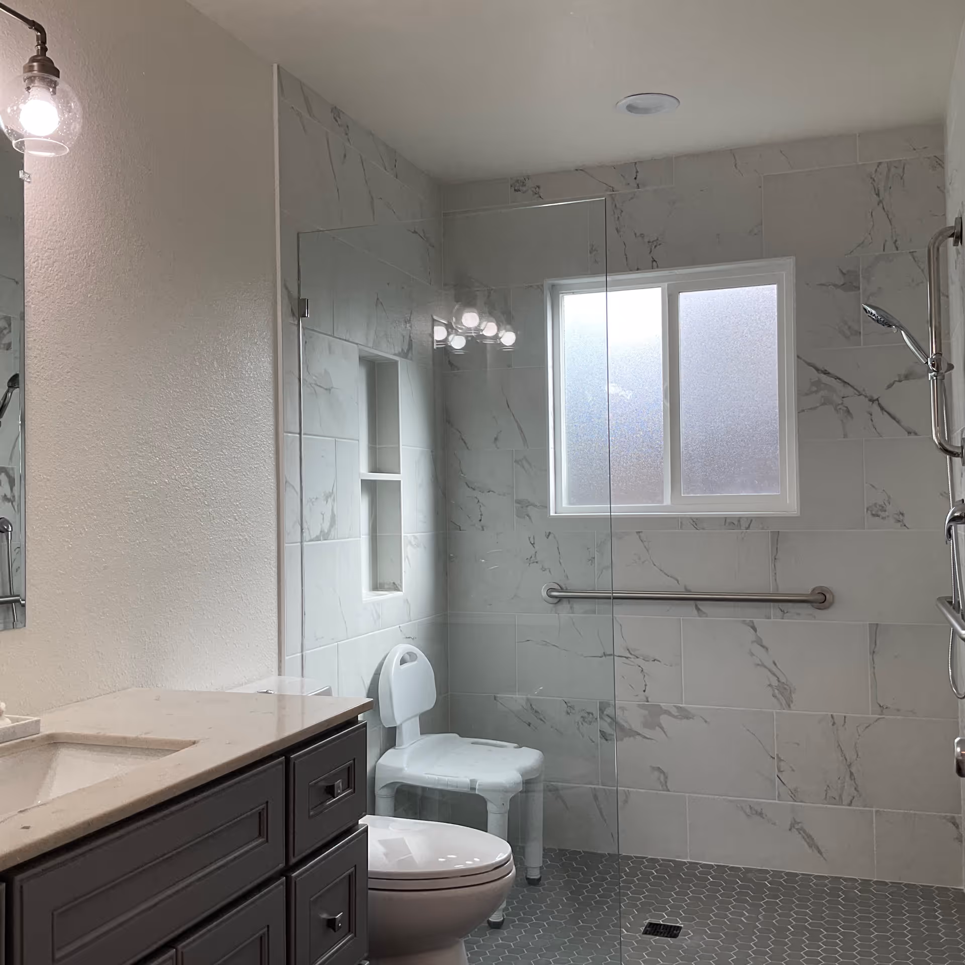 A modern bathroom with marble-patterned tiles on the walls and hexagonal gray tiles on the floor. There is a white toilet and a white shower chair inside a walk-in shower area with a glass partition. A grab bar is mounted on the wall under a frosted window. A vanity with a beige countertop and dark cabinetry is visible on the left side, along with a wall-mounted light fixture above the mirror.