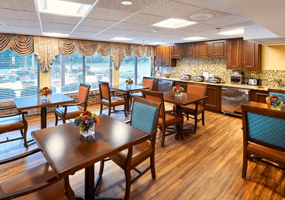 A bright dining area with wooden tables and chairs, each table decorated with a small vase of colorful flowers. Large windows with ornate valances allow natural light to fill the room. The back wall features a kitchen area with wooden cabinets, a mosaic tile backsplash, and stainless steel appliances including a dishwasher, toaster, and chafing dishes.