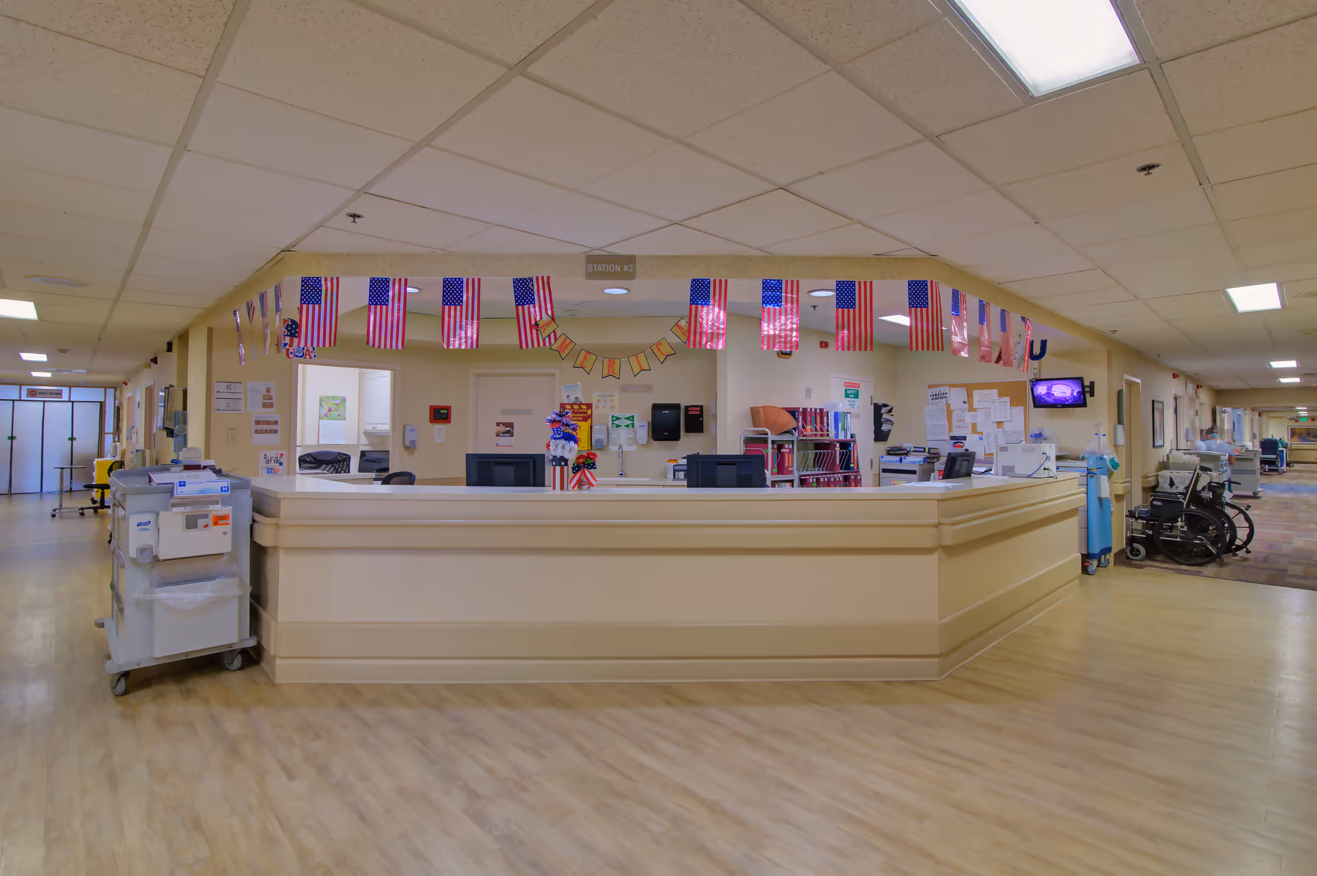 A nursing station in a senior living facility decorated with small American flags and a 'Happy Fourth' banner. The station has a beige counter with office equipment, chairs, and medical carts nearby. The hallway extends on both sides with additional medical equipment and wheelchairs visible.