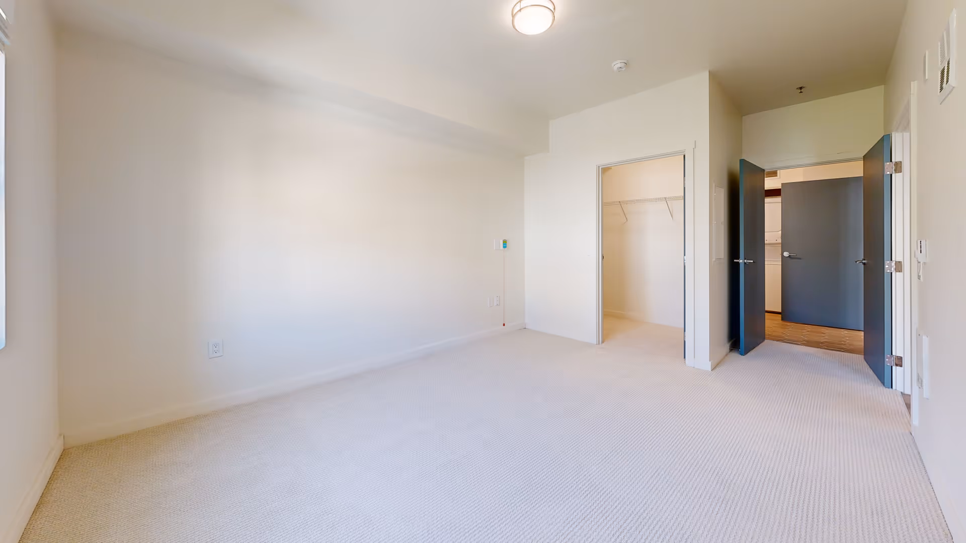 Empty bedroom with beige carpet, white walls, a ceiling light, an open closet with wire shelving, and two open dark-colored doors leading to a hallway and laundry area.