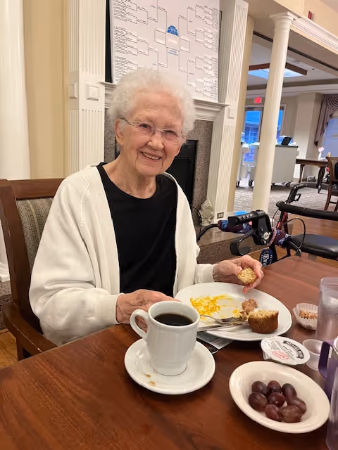 An elderly woman smiles at a dining table with a plate of muffins, coffee, and grapes in a senior living dining room.