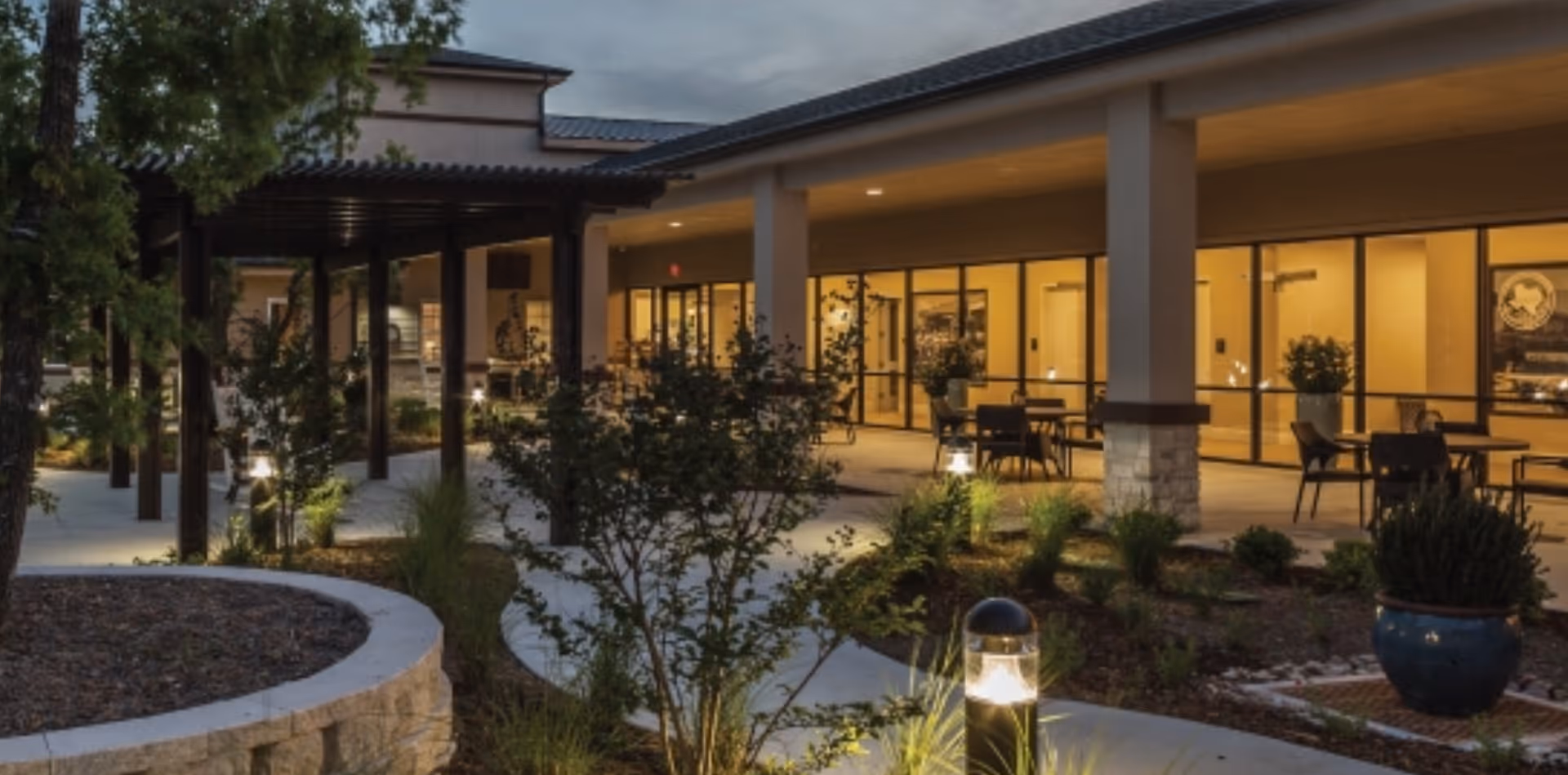 Outdoor patio area at dusk with a covered walkway, landscaped garden beds, and seating tables near large glass windows of a building.