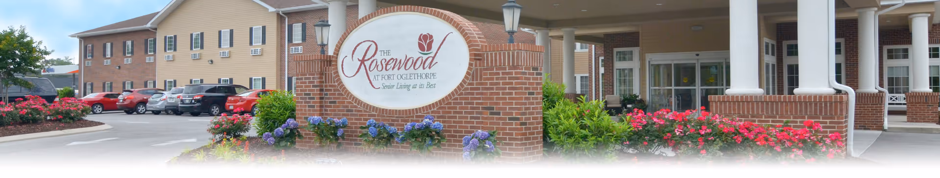 Front entrance of The Rosewood at Fort Oglethorpe senior living building with a brick sign, landscaped flowers, and parked cars in the driveway.