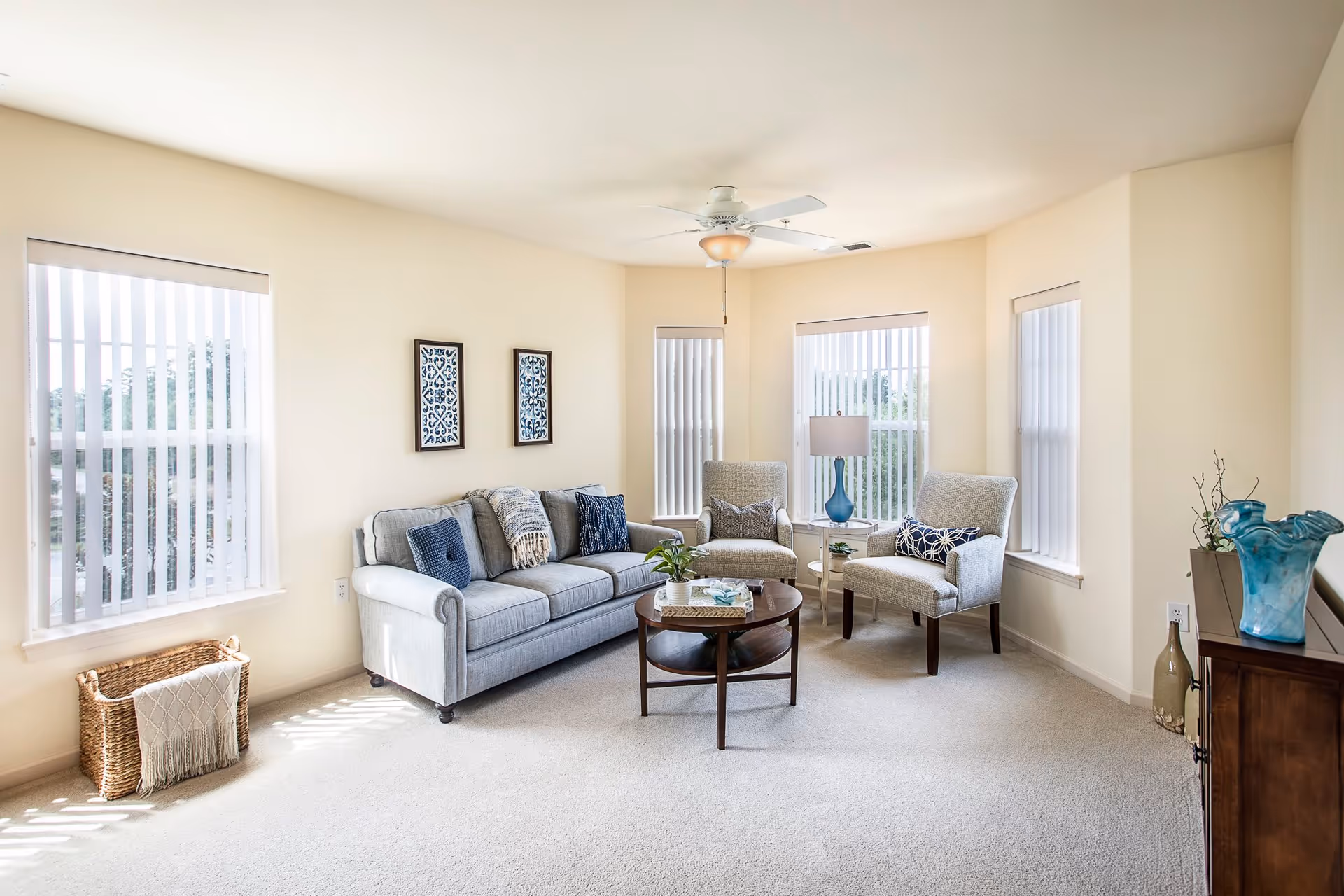 A bright living room with beige walls and carpet, featuring a light gray sofa with blue and patterned cushions, two beige armchairs with pillows, a round wooden coffee table with decorative items, a blue table lamp on a small side table, three tall windows with vertical blinds, and a wooden cabinet with blue glass vases and decorative bottles.