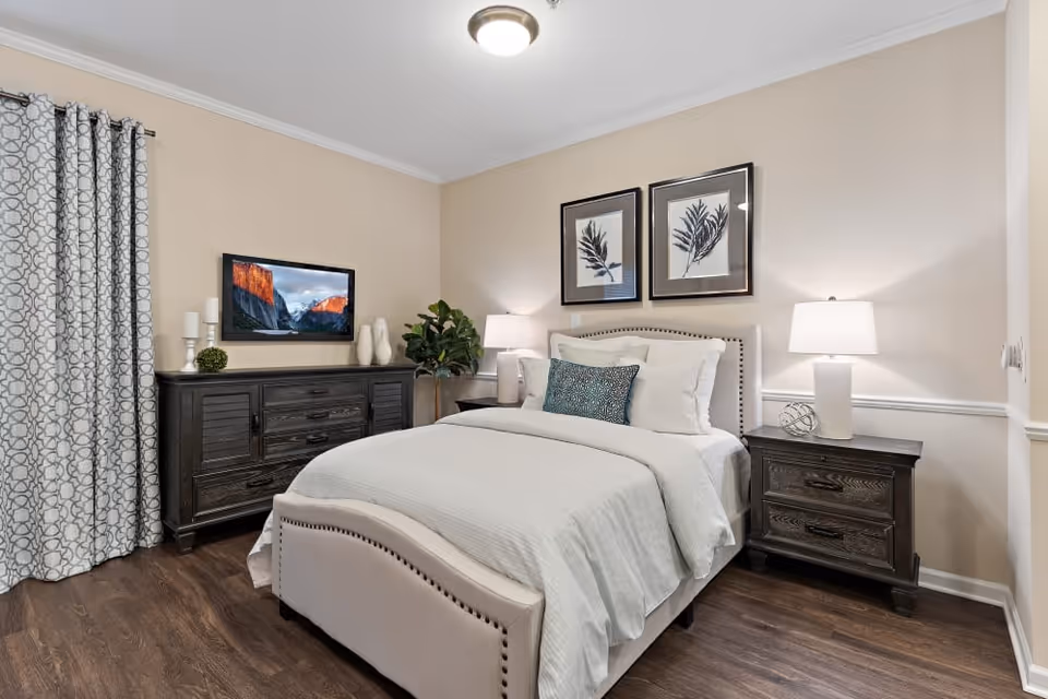 A neatly made bed with white bedding and decorative pillows in a bedroom. The room features two dark wooden nightstands with white lamps, a dark wooden dresser with decorative items and a mounted flat-screen TV above it. There are patterned curtains on the left side and two framed botanical prints hanging above the bed. The floor is wooden and the walls are painted beige.