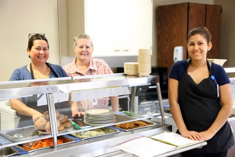 Three women standing behind a cafeteria-style food serving counter with trays of food including carrots, mashed potatoes, and bread rolls. Two women are behind the counter smiling, and one woman wearing a black apron stands in front of the counter smiling. Plates and bowls are stacked on the counter.