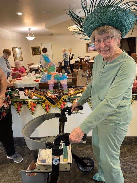 An elderly woman wearing a green outfit and a large green hat stands with a walker in a communal dining area decorated with colorful banners and a small piñata on the table. Other elderly people are seated and standing in the background, engaging in conversation.