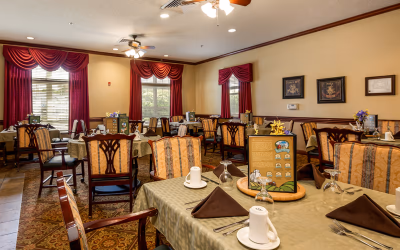A dining room with multiple tables set for a meal, featuring beige tablecloths, brown napkins, white cups, and glassware. The room has patterned carpet, wooden chairs with upholstered seats and backs, and windows with red curtains. Ceiling fans and framed artwork are visible on the walls.