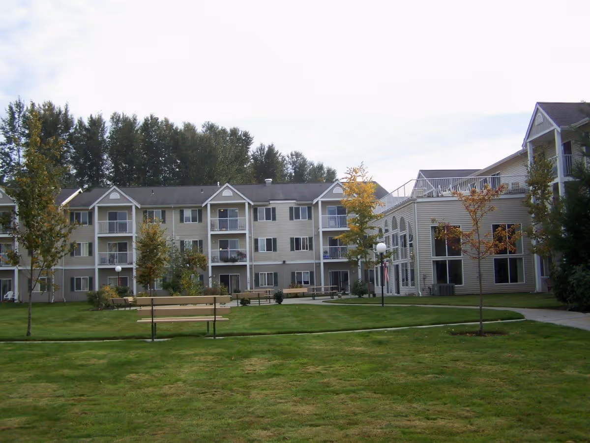 Exterior view of The Vintage at Mount Vernon senior living facility showing a three-story building with balconies, surrounded by a well-maintained lawn with benches and young trees.