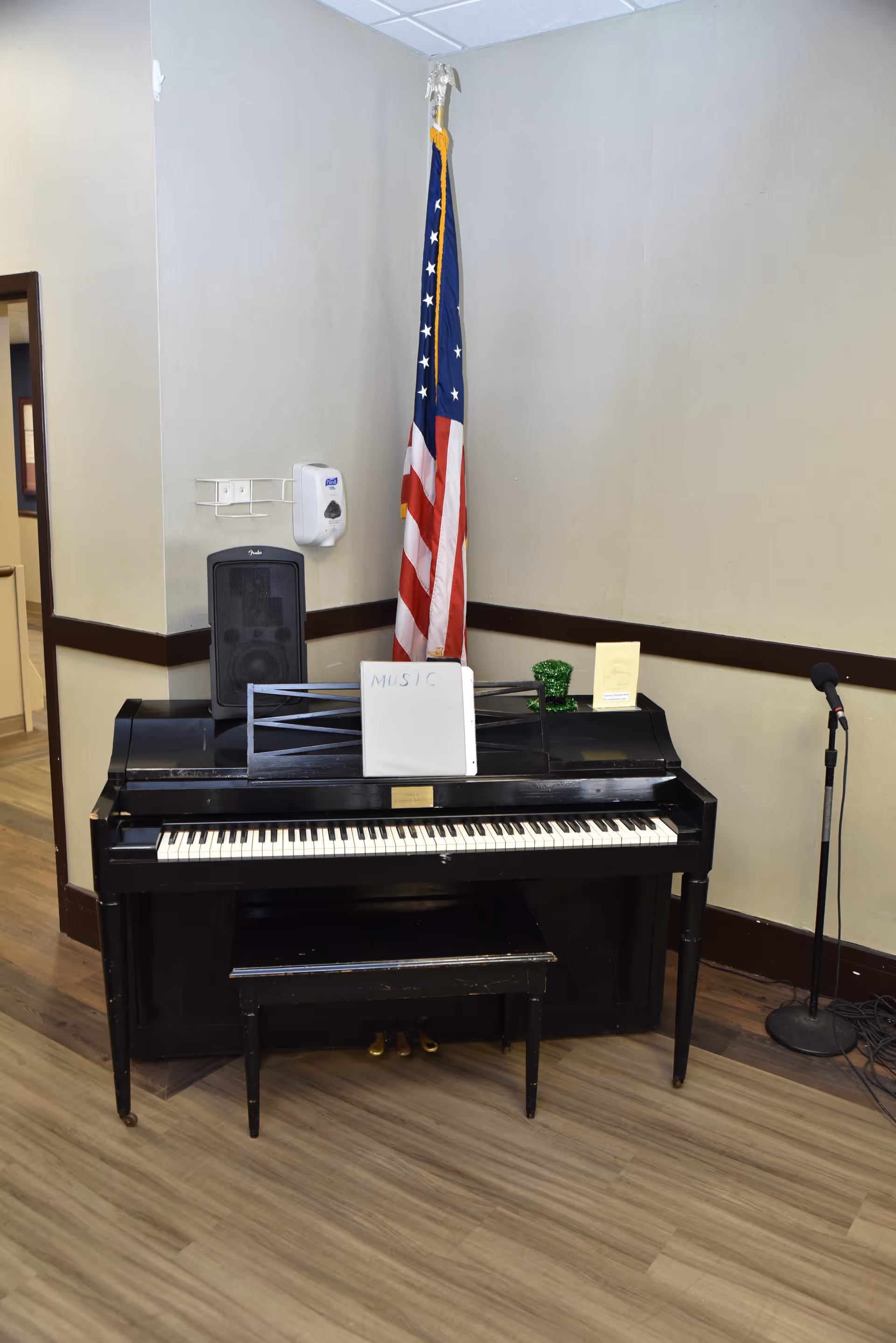 A black upright piano with a matching bench in a corner of a room. On top of the piano is a music stand holding a sheet labeled 'MUSIC', a small green decorative item, and a yellow booklet. Behind the piano is an American flag standing in the corner. To the right of the piano is a microphone on a stand. The room has light-colored walls with dark trim and wood-patterned flooring.