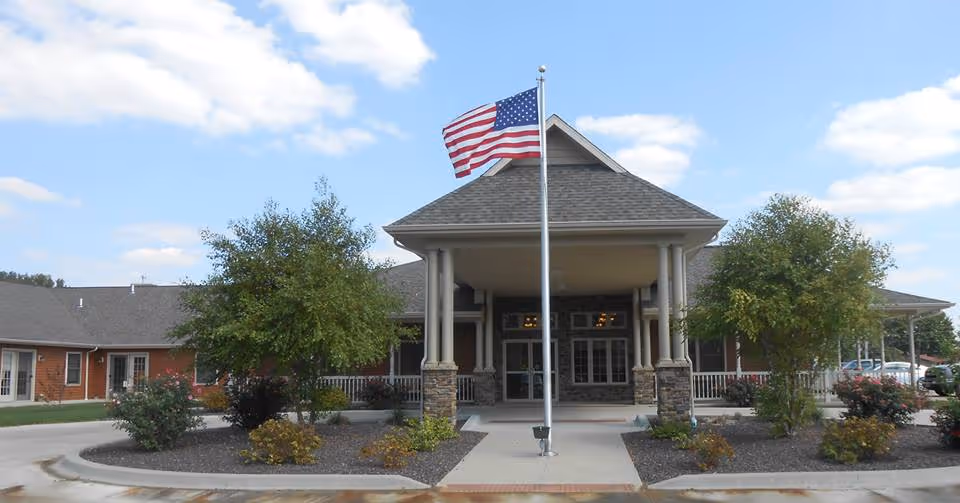 Front exterior view of Timber Creek Village Assisted Living of Robinson building with a covered entrance supported by columns, an American flag on a flagpole in front, trees and shrubs on either side, and a partly cloudy sky.