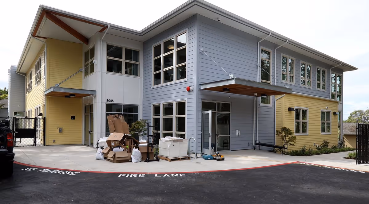 Front exterior of a two-story modern senior living building with yellow and gray siding, large windows, an entrance canopy, and boxes on the driveway.