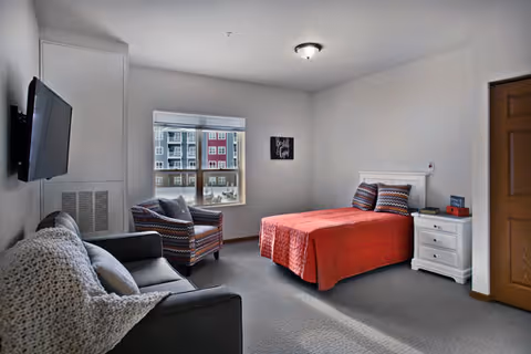 A cozy bedroom with a bed covered in a red quilt, two pillows, a white nightstand with a lamp and books, a patterned armchair near a window showing an outside view of a building, a gray sofa with a knitted throw blanket, and a wall-mounted TV.