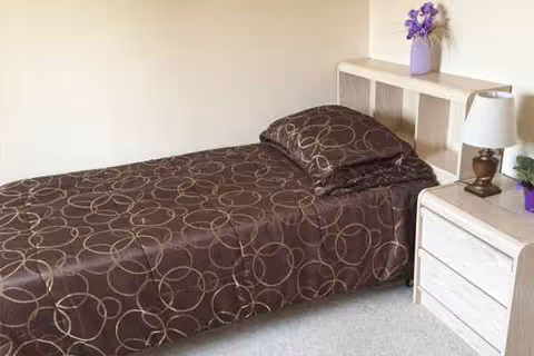 A small bedroom with a single bed, nightstand, lamp, and purple flowers.