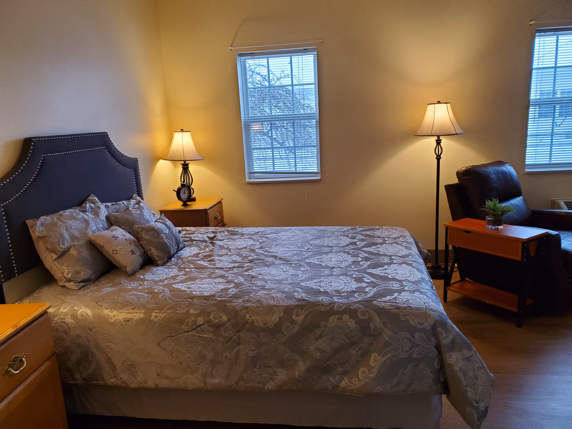 A cozy bedroom with a large bed covered in a patterned bedspread and multiple pillows. There are two windows with blinds, a nightstand with a lamp and an alarm clock on the left side of the bed, and a floor lamp next to a dark armchair with a small wooden side table holding a small plant.