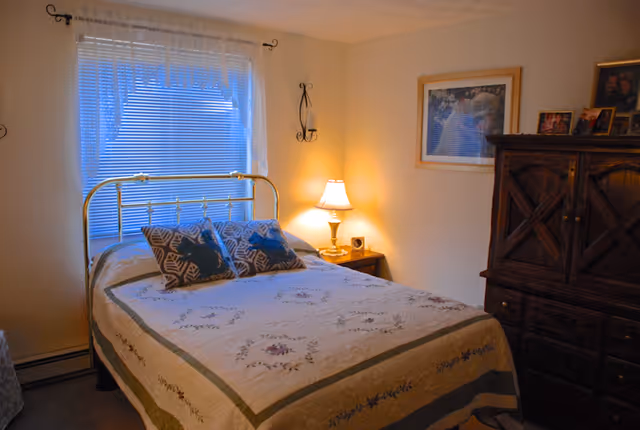 A cozy bedroom with a brass bed frame, floral quilt, and two patterned pillows. A window with blinds and sheer curtains is behind the bed. A wooden nightstand with a lit table lamp is beside the bed. On the right side, there is a dark wooden dresser with framed photos on top and a framed picture hanging on the wall above it.