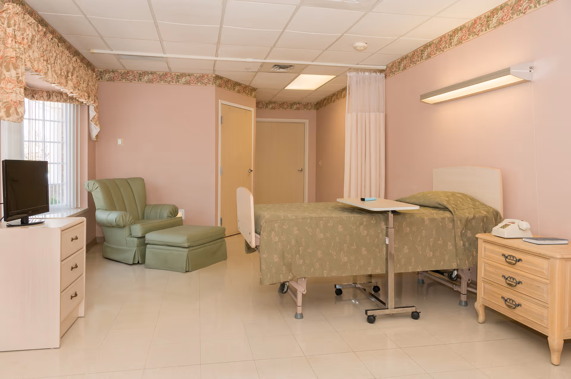A senior living facility room with a hospital-style bed covered in a green patterned bedspread, a green upholstered armchair with matching ottoman near a window with floral curtains, a small wooden dresser with a TV on top, and a bedside table with a telephone and book. The walls are painted light pink with floral wallpaper border near the ceiling.