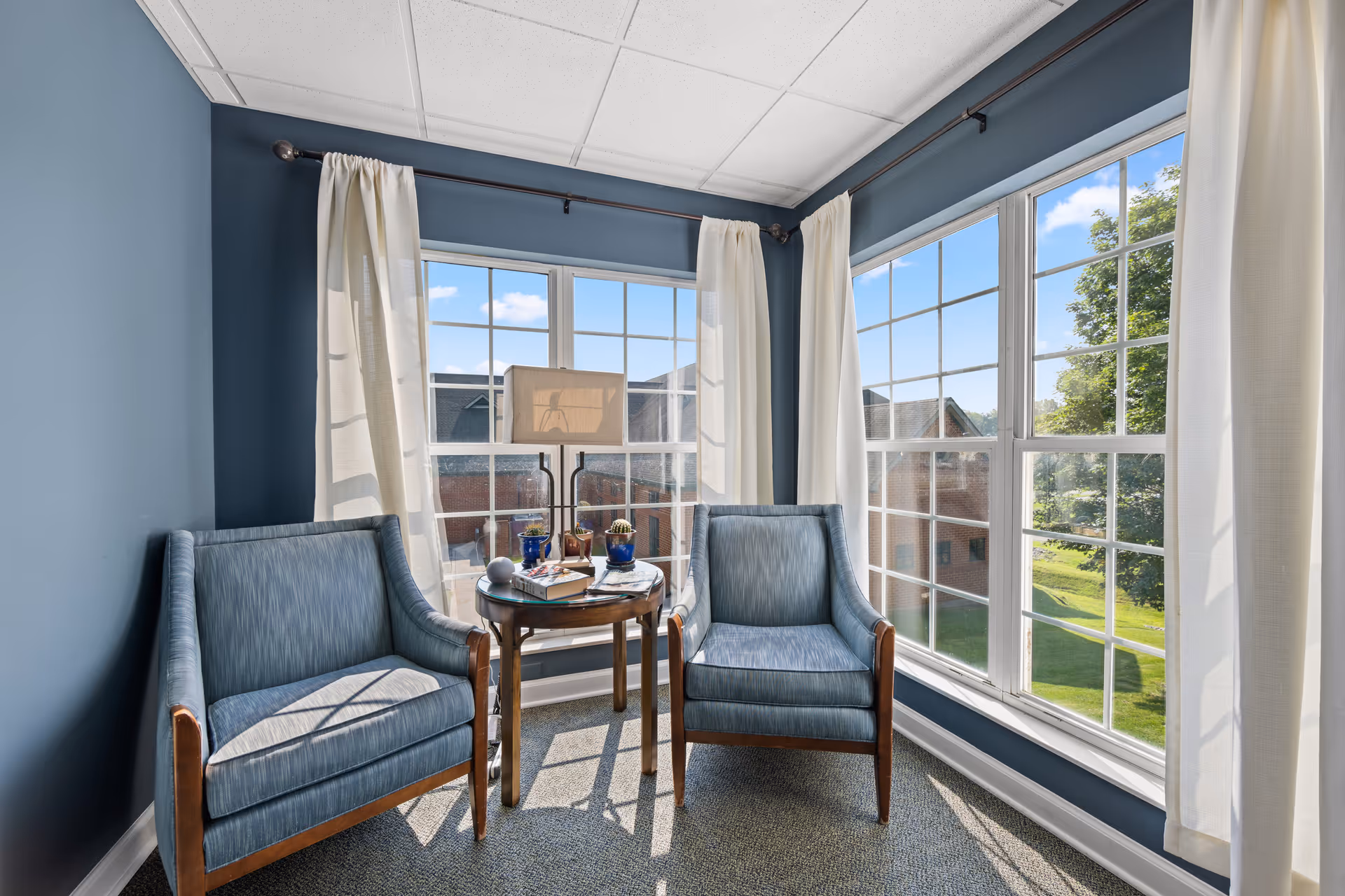 Sunlit seating area with two blue armchairs flanking a small round table in a corner with large grid windows and white curtains.