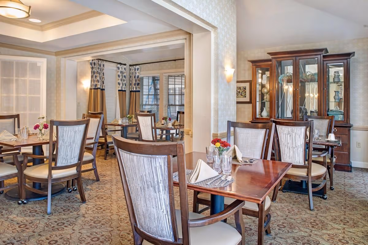 Bright, upscale dining room with wooden tables and chairs set with napkins and glassware and a china cabinet in the background.