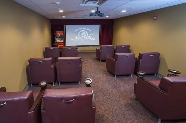 A small theater room with several rows of comfortable maroon armchairs facing a large screen displaying the Metro Goldwyn Mayer logo. There is a popcorn machine in the back corner of the room. The walls are painted beige and the ceiling has recessed lighting.
