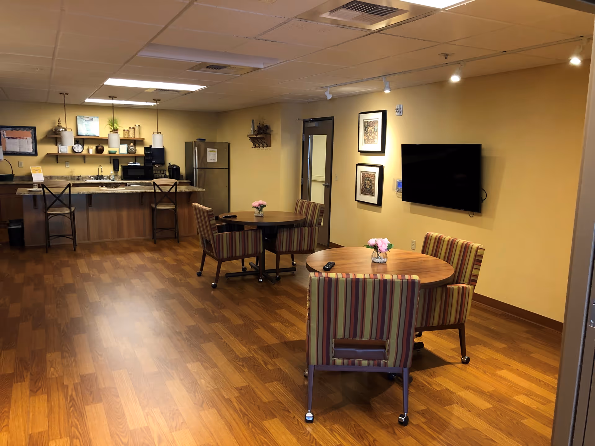 A cozy common area with wooden flooring featuring two round tables with striped cushioned chairs on wheels. Each table has a small vase with pink flowers. On the right wall, there is a mounted flat-screen TV and two framed artworks. In the background, there is a kitchenette with a granite countertop, two pendant lights, a refrigerator, microwave, and various kitchen items on shelves. The walls are painted a warm yellow color.